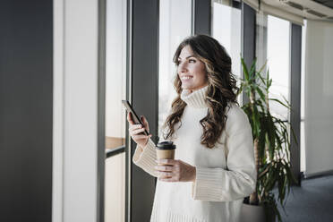 Smiling businesswoman with smart phone holding disposable cup near window in office - EBBF05390