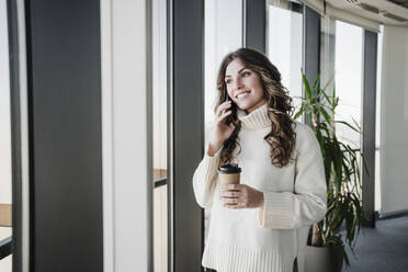 Businesswoman with disposable cup talking on mobile phone near window in office - EBBF05391
