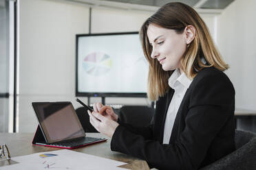 Lächelnde Geschäftsfrau, die ein Smartphone neben einem Tablet-PC im Büro benutzt - EBBF05405