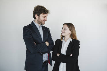 Smiling businesswoman and businessman standing with arms crossed looking at each other in front of wall - EBBF05420