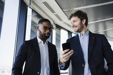 Smiling businessman with smart phone looking at colleague talking in office - EBBF05453