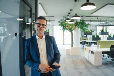 Businessman with smart phone leaning on wall at office - JOSEF06958