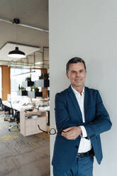 Working man holding eyeglasses leaning on wall at office - JOSEF06967