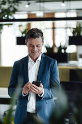 Smiling businessman using smart phone standing at office - JOSEF06971