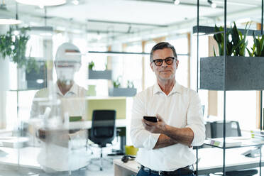 Lächelnder Geschäftsmann mit Smartphone, der in der Nähe eines Regals im Büro steht - JOSEF06982