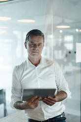 Businessman holding tablet PC leaning on glass wall at office - JOSEF06986