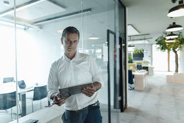 Selbstbewusster Geschäftsmann mit Tablet-PC, der an einer Glaswand im Büro lehnt - JOSEF06988