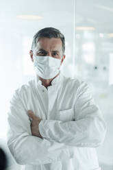 Scientist with protective face mask leaning on glass wall at medical clinic - JOSEF06991