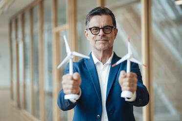 Smiling businessman holding wind turbines in hand at office - JOSEF07001