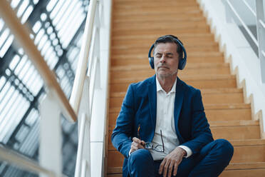 Businessman with eyes closed listening music on headphones at office - JOSEF07006