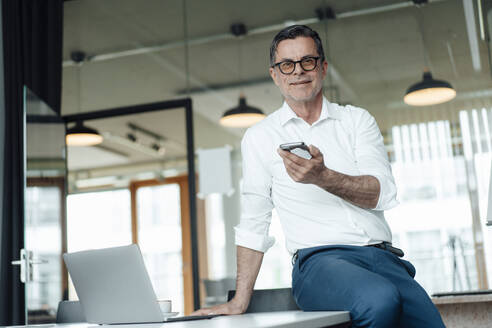 Smiling businessman with smart phone sitting on table at office - JOSEF07010
