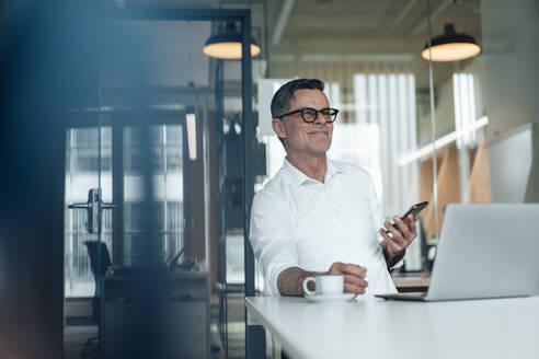 Lächelnder Geschäftsmann mit Smartphone träumt im Büro - JOSEF07012