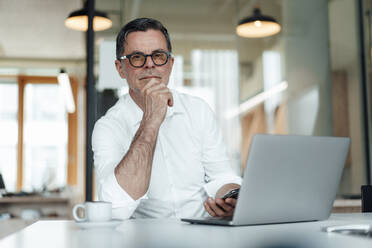 Selbstbewusster Geschäftsmann mit Hand am Kinn, der mit einem Laptop auf dem Tisch im Büro sitzt - JOSEF07013