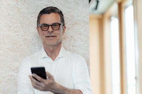 Businessman with eyeglasses holding smart phone in front of wall - JOSEF07015