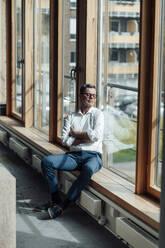 Geschäftsmann mit verschränkten Armen sitzt auf der Fensterbank im Büro an einem sonnigen Tag - JOSEF07018