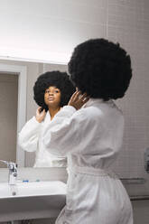 Junge Frau mit Hand in den Haaren schaut in den Spiegel in der Wohnung - PNAF02993