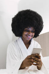 Afro-Frau im Bademantel mit Smartphone im Schlafzimmer zu Hause - PNAF03011