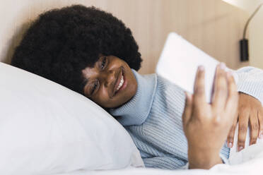 Smiling Afro woman using smart phone on bed at home - PNAF03031
