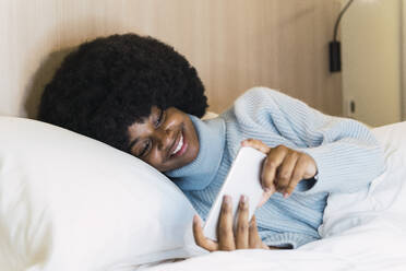 Smiling woman with turtleneck sweater watching on smart phone in bedroom at home - PNAF03032