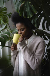 Afro woman drinking coffee by monstera plants at home - VPIF05264