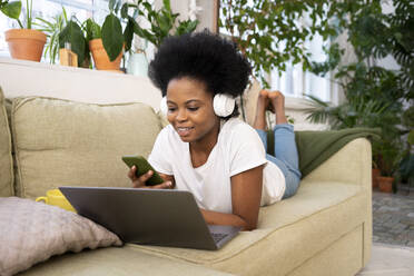 Geschäftsfrau mit Kopfhörern und Laptop, die auf dem Sofa zu Hause ihr Smartphone benutzt - VPIF05297