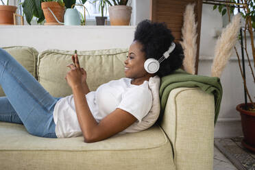 Smiling Afro woman with headphones using smart phone on sofa in living room - VPIF05302