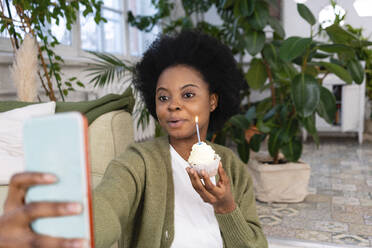 Afro woman taking selfie with cupcake in living room at home - VPIF05316