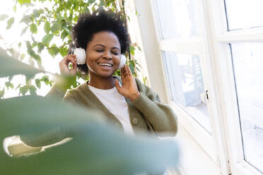 Glückliche Afro-Frau, die zu Hause am Fenster Musik über Kopfhörer hört - VPIF05332