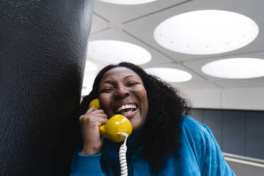 Glückliche Frau, die in einen altmodischen gelben Telefonhörer spricht, der an einer Säule lehnt - ASGF02106