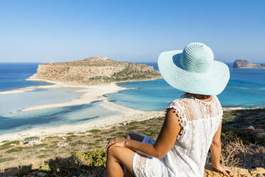 Porträt einer Frau mit Hut, die auf einer Anhöhe sitzend den idyllischen Strand und die Lagune bewundert, Balos, Kreta, Griechische Inseln, Griechenland, Europa - RHPLF21407