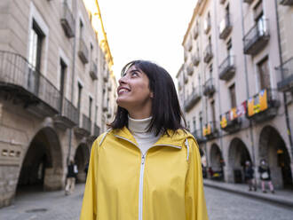 Lächelnde Frau in gelbem Regenmantel auf dem Fußweg in der Stadt - AMRF00002