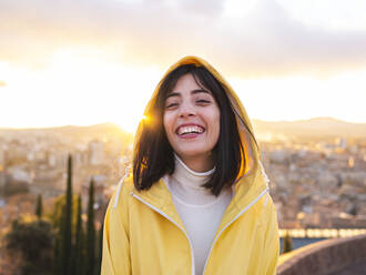 Lächelnde junge Frau in gelbem Regenmantel mit Stadt im Hintergrund bei Sonnenuntergang - AMRF00003