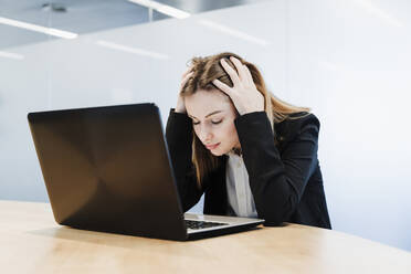 Erschöpfte Geschäftsfrau mit dem Kopf in den Händen sitzt mit Laptop am Tisch im Büro - EBBF05461