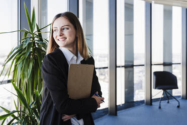 Happy businesswoman holding clipboard at office - EBBF05465