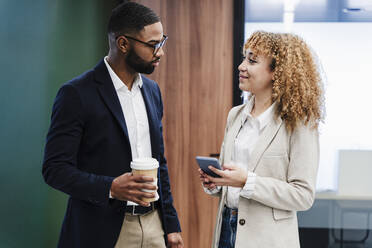 Smiling businesswoman with smart phone standing with colleague holding disposable cup at coworking office - EBBF05478