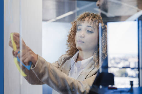 Businesswoman sticking adhesive note on glass wall at office - EBBF05491