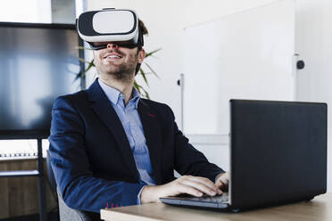Businessman with laptop wearing virtual reality headset in office - EBBF05503