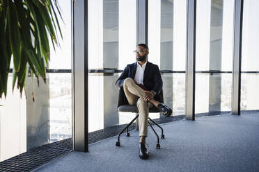 Businessman sitting on chair near glass window in office - EBBF05522