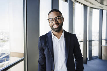 Happy businessman leaning on window in office - EBBF05527