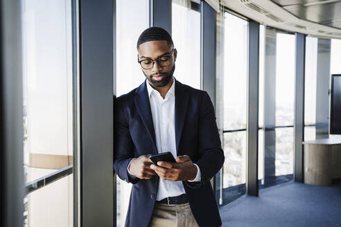 Working man using smart phone leaning on window in office - EBBF05528