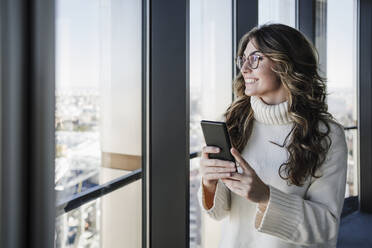Glückliche Geschäftsfrau hält Smartphone in der Nähe des Fensters im Büro - EBBF05562