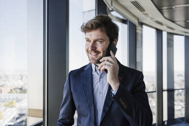 Lächelnder Geschäftsmann, der im Büro mit seinem Handy telefoniert - EBBF05568