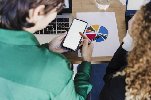 Geschäftsfrau mit Stift und Smartphone am Schreibtisch eines Kollegen in einem Coworking-Büro - EBBF05583