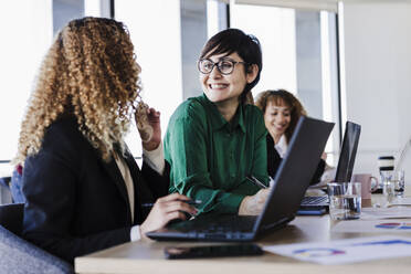 Happy businesswoman looking at young colleague with laptop in coworking office - EBBF05586