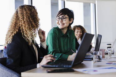 Zufriedene Geschäftsfrau mit Blick auf einen jungen Kollegen mit Laptop in einem Coworking-Büro - EBBF05586