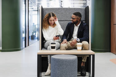 Junger Geschäftsmann im Gespräch mit einem Kollegen, der sein Smartphone in der Kaffeepause in einem Coworking-Büro benutzt - EBBF05603