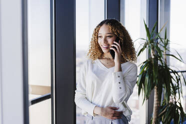 Junge Geschäftsfrau mit lockigem Haar, die in der Nähe eines Fensters im Büro mit einem Smartphone spricht - EBBF05616