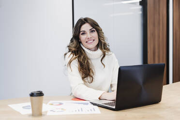Lächelnde Geschäftsfrau sitzt am Schreibtisch mit Laptop im Büro - EBBF05618