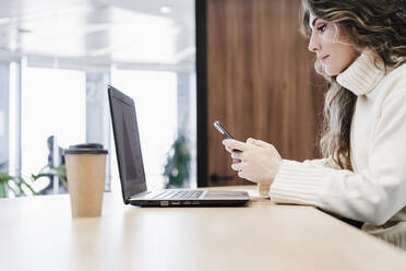 Geschäftsfrau benutzt Smartphone über Laptop am Schreibtisch im Büro - EBBF05620