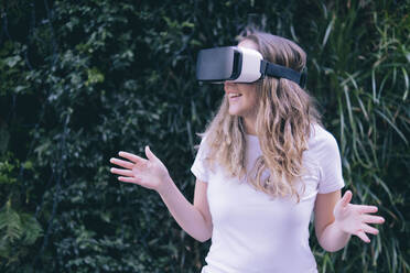 Frau gestikuliert mit Virtual-Reality-Headset vor einer Hecke - AMWF00170